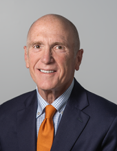 Jim is smiling and wearing a blue suit jacket, blue and white plaid shirt and orange tie.