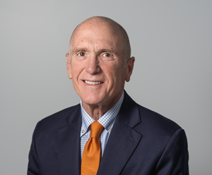 Jim is smiling and wearing a blue suit jacket, blue and white plaid shirt and orange tie.